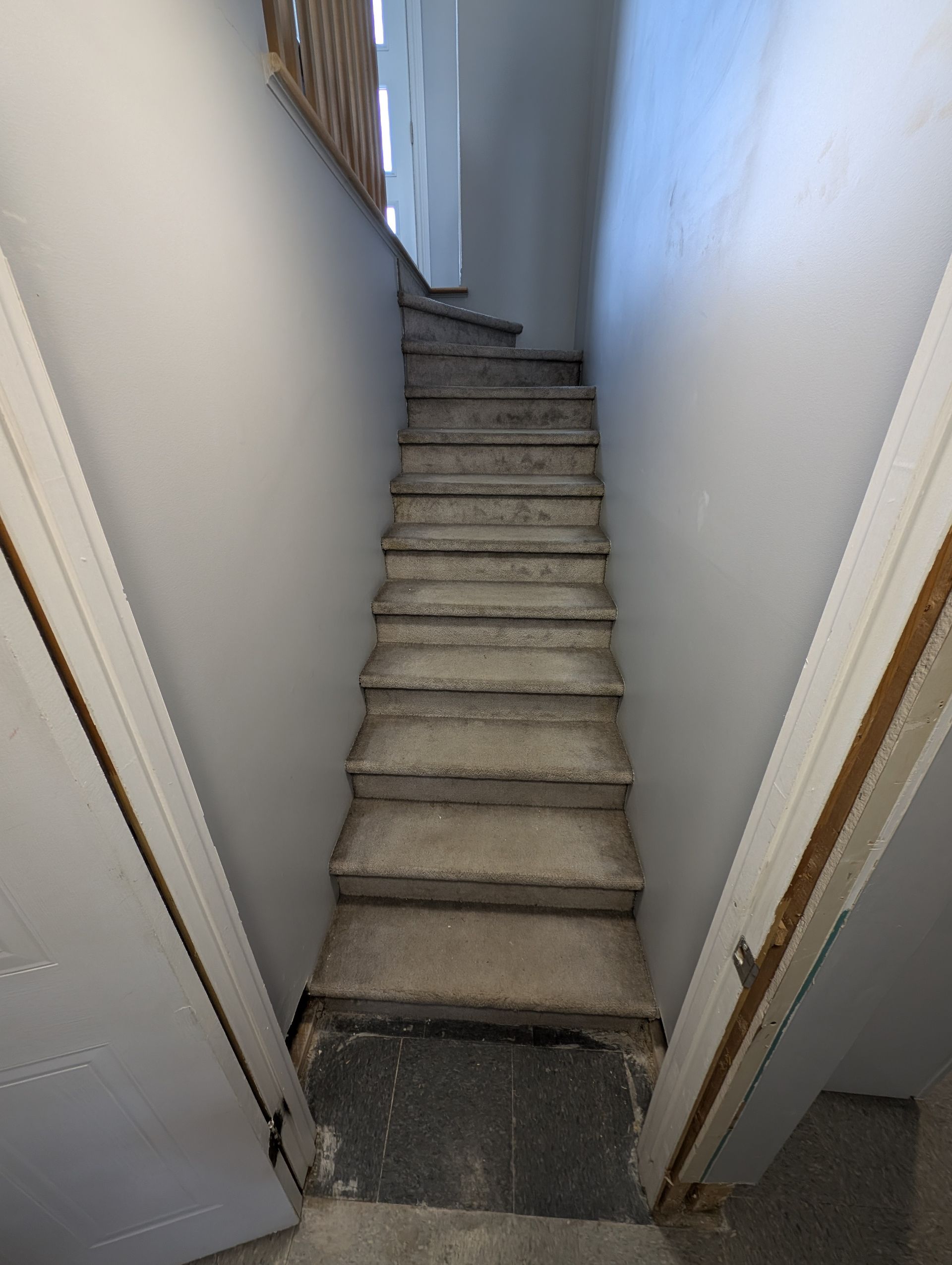 Un escalier étroit recouvert de moquette, entre des murs blancs, mène à un palier avec une rampe en bois.