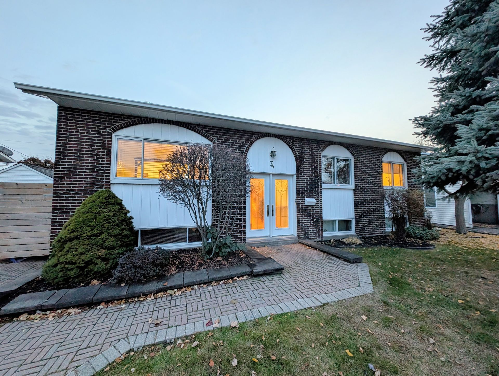 Maison moderne en briques de deux étages au crépuscule, avec des fenêtres éclairées et une allée pavée menant à la porte d'entrée.