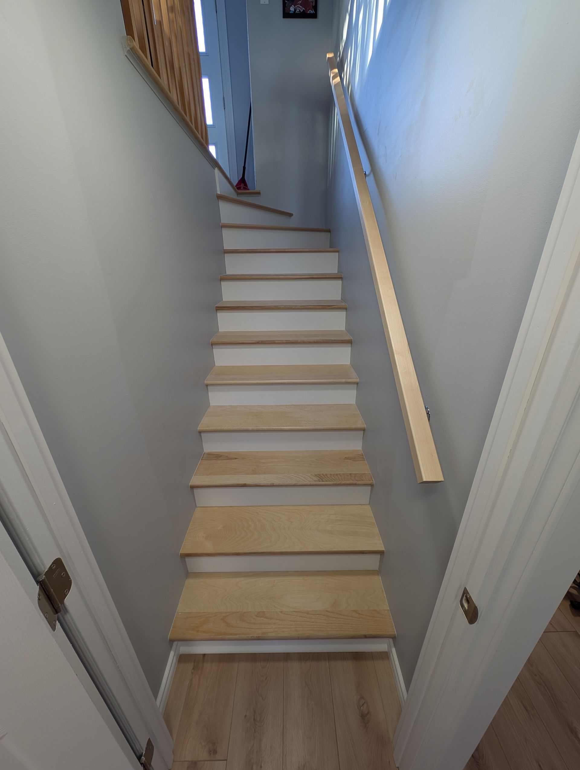 Escalier intérieur étroit aux murs blancs, aux marches en bois clair et à la rampe, vu d'en bas.