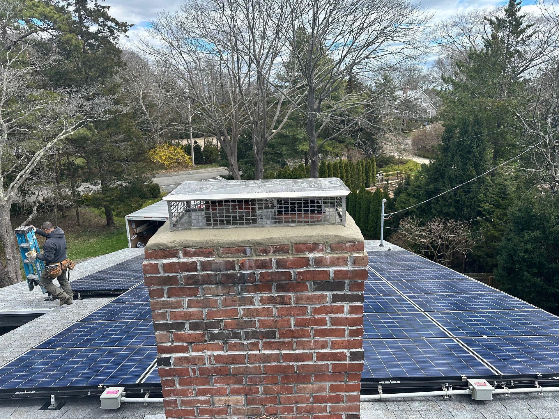 Chimney on rooftop with solar panels and two workers; trees in background.