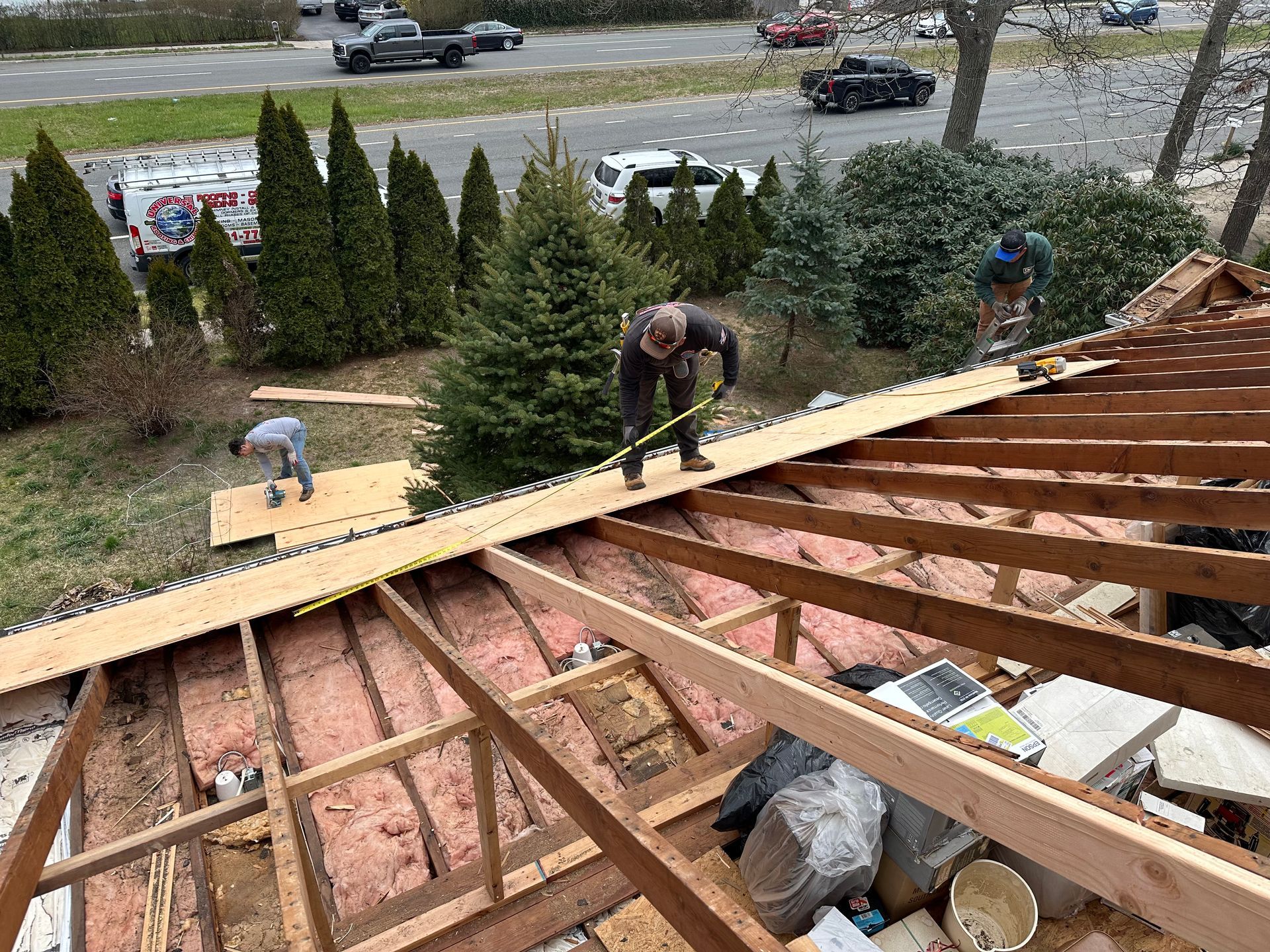 Construction workers repair a roof, measuring and placing wood planks.  Outdoor setting with vehicles in the background.