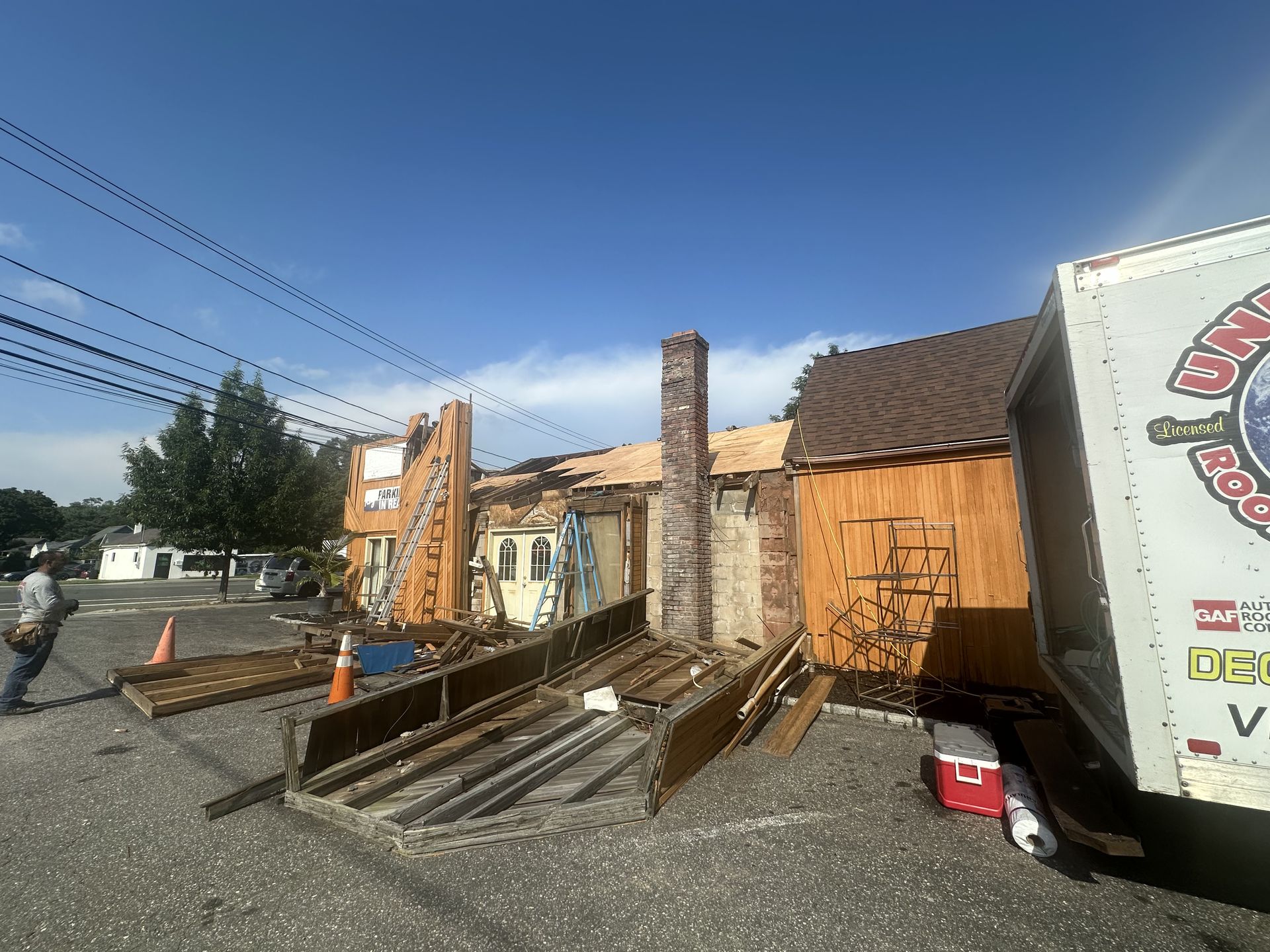Building heavily damaged by fire, partially collapsed. Debris and a chimney are visible under a bright blue sky.