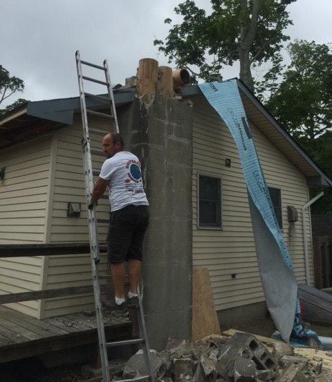 Man on ladder working on a chimney on a house with a damaged roof under an overcast sky.