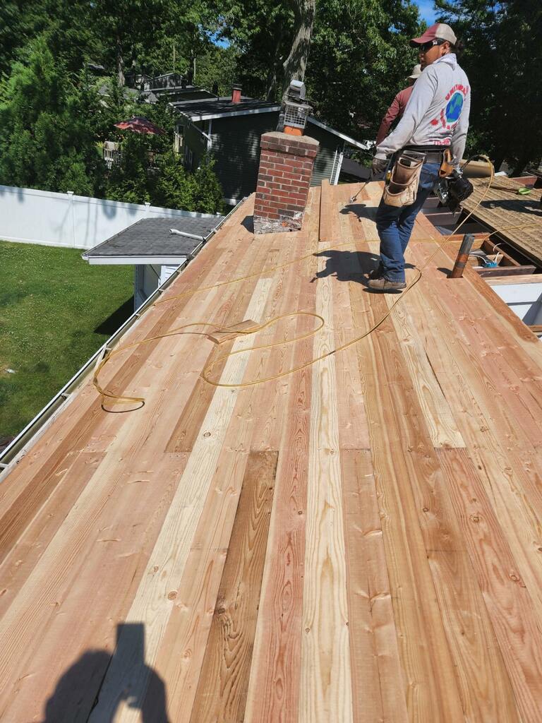 Roofer standing on a new cedar shake roof, near a brick chimney, holding a tool.