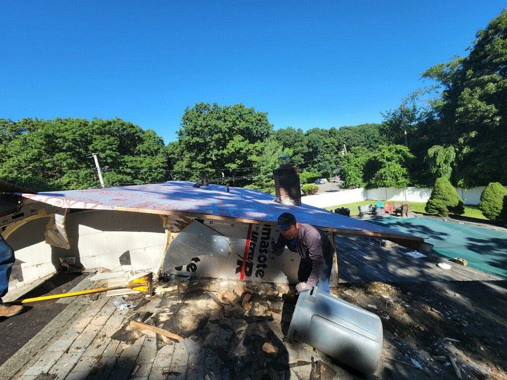 Roof being repaired; a worker is cutting a piece of metal. Bright blue sky, green trees in the background.