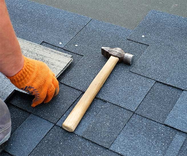 Person installing dark blue asphalt shingles on a roof with a hammer, wearing orange gloves.