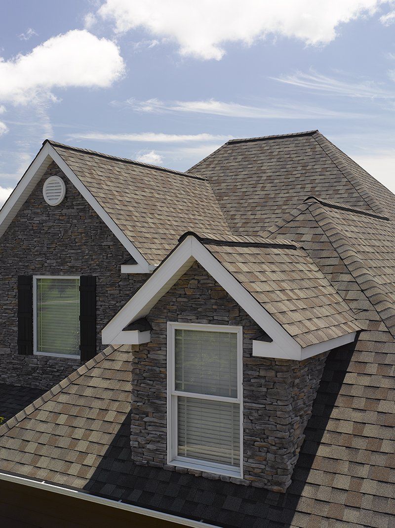 House exterior with stone siding, multiple dormers, and a shingle roof against a blue sky.