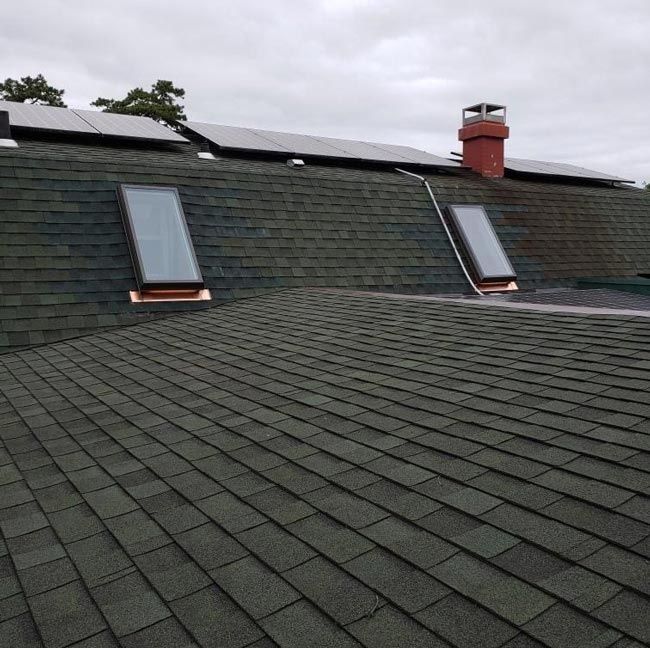 A dark green shingled roof with solar panels, skylights, and a chimney against a cloudy sky.
