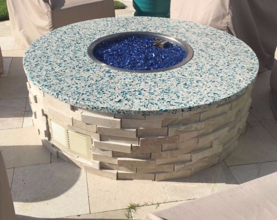 Round fire pit with blue glass and light stone facade on a patio.