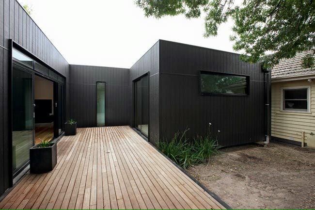 Modern black-clad building with wood deck patio; large windows, rectangular shape, greenery.