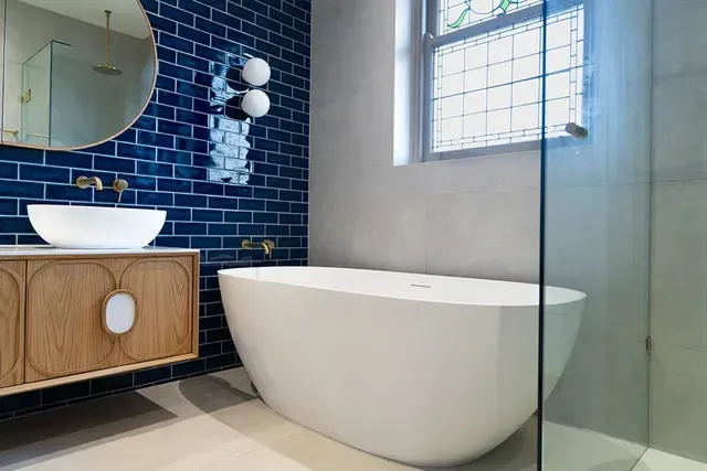 Modern bathroom with blue tile, a white tub, wooden vanity, and a stained glass window.