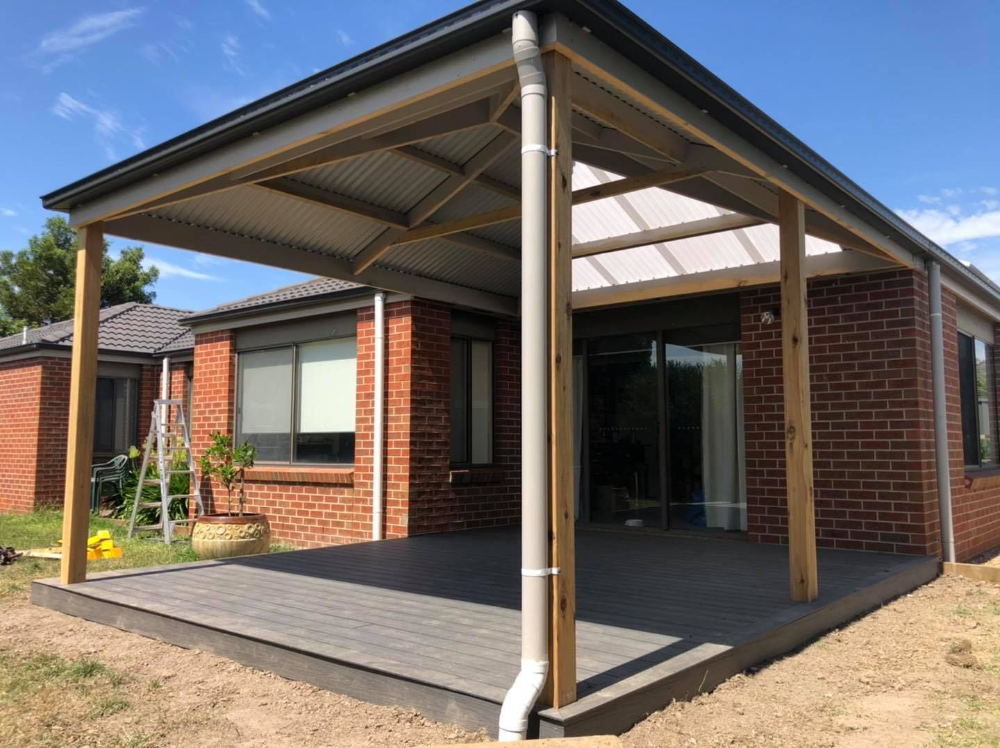 Patio with brick house, gray paving, wooden frame, and corrugated roof. Bright day, blue sky.