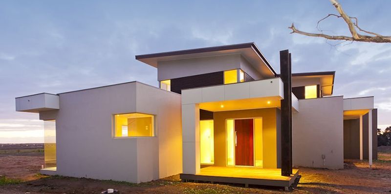 Modern, white rectangular house with brown door, illuminated by warm lights at dusk.
