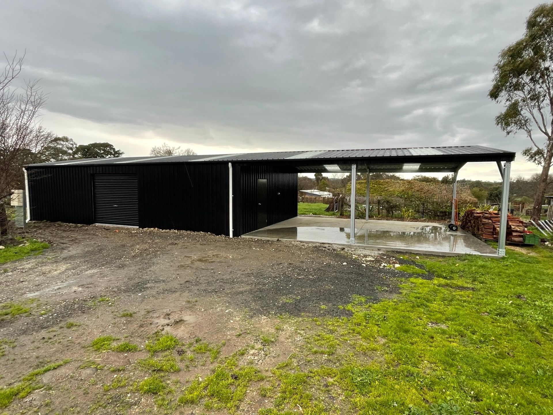 Black shed and carport on a gravel driveway, against an overcast sky.