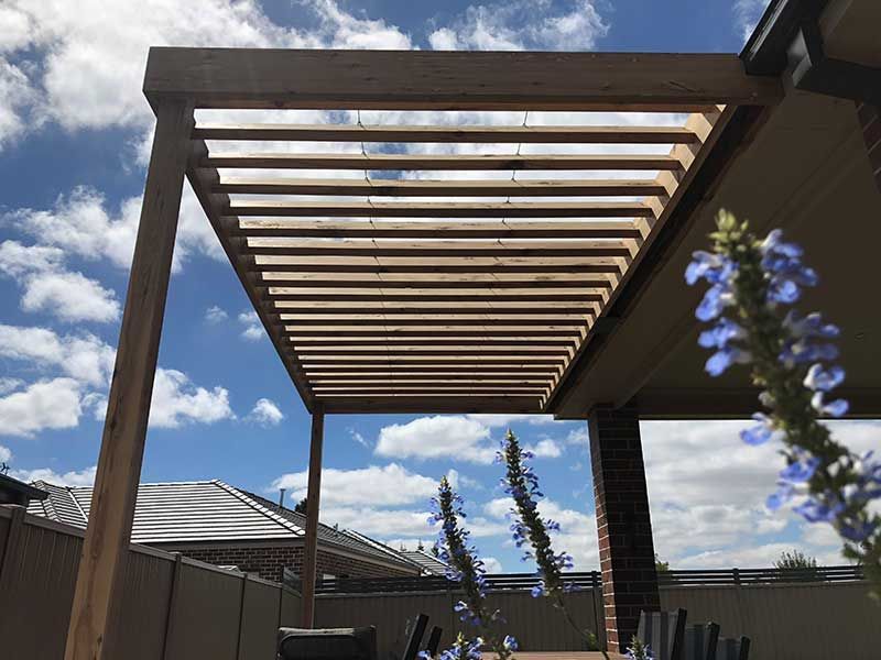 Wooden pergola with horizontal slats against a cloudy blue sky, near a covered patio and blooming flowers.