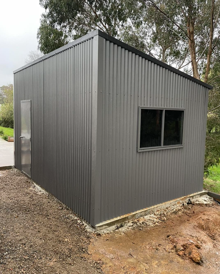 Gray corrugated metal shed with a window and door, on a concrete base outdoors.