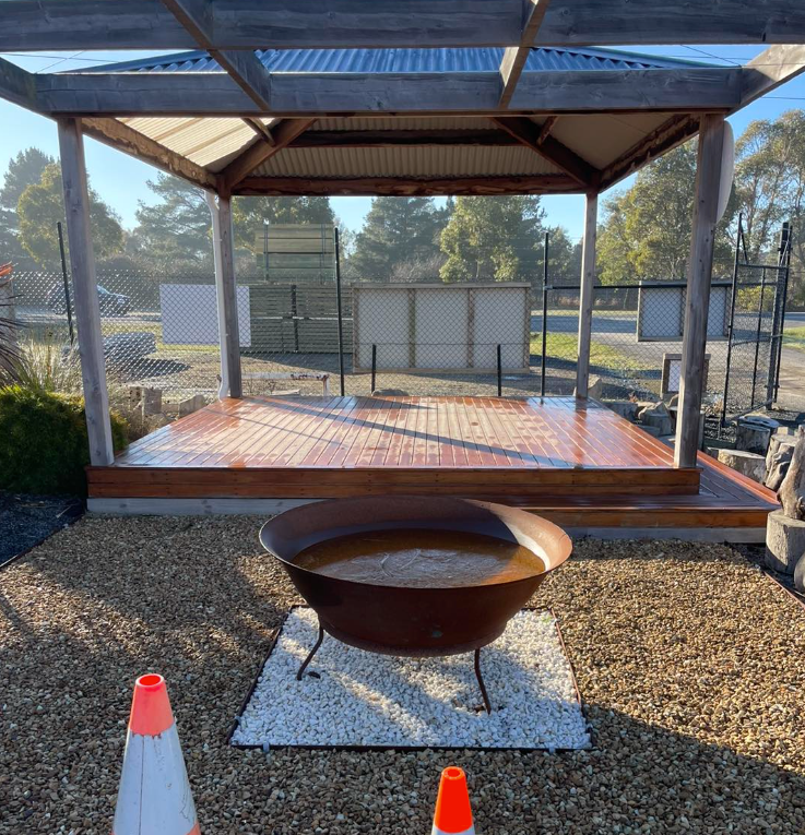 Gazebo with wooden deck, fire pit on gravel, orange traffic cones, and fence in background.