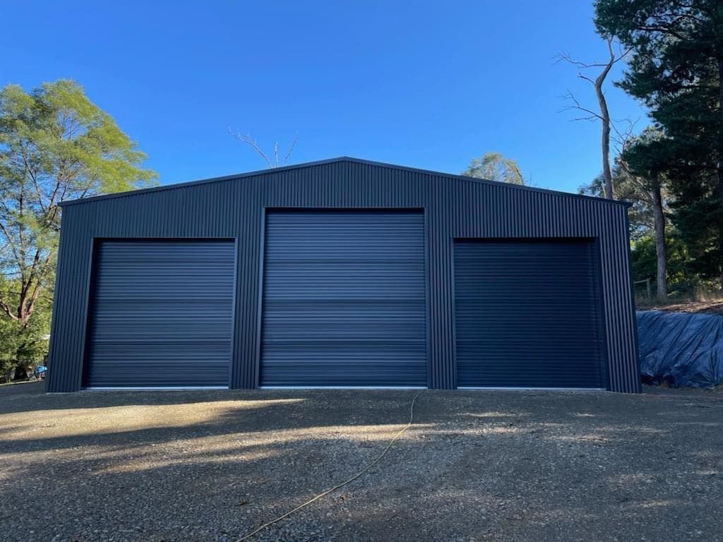 Dark gray metal shed with three garage doors on a gravel lot.