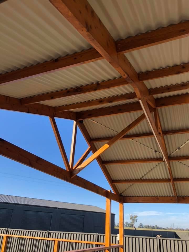 Wooden-framed pergola roof with corrugated metal panels, supported by angled beams. Bright sky background.