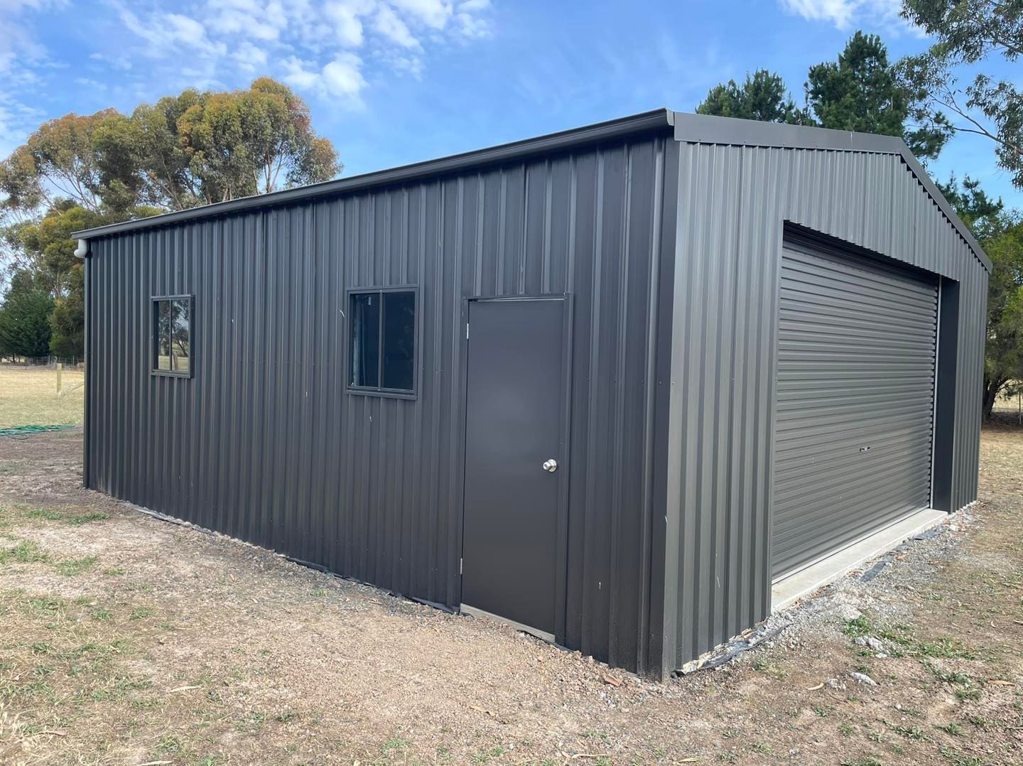 Dark gray corrugated metal shed with a roll-up door, door, and two windows on a grassy lot.