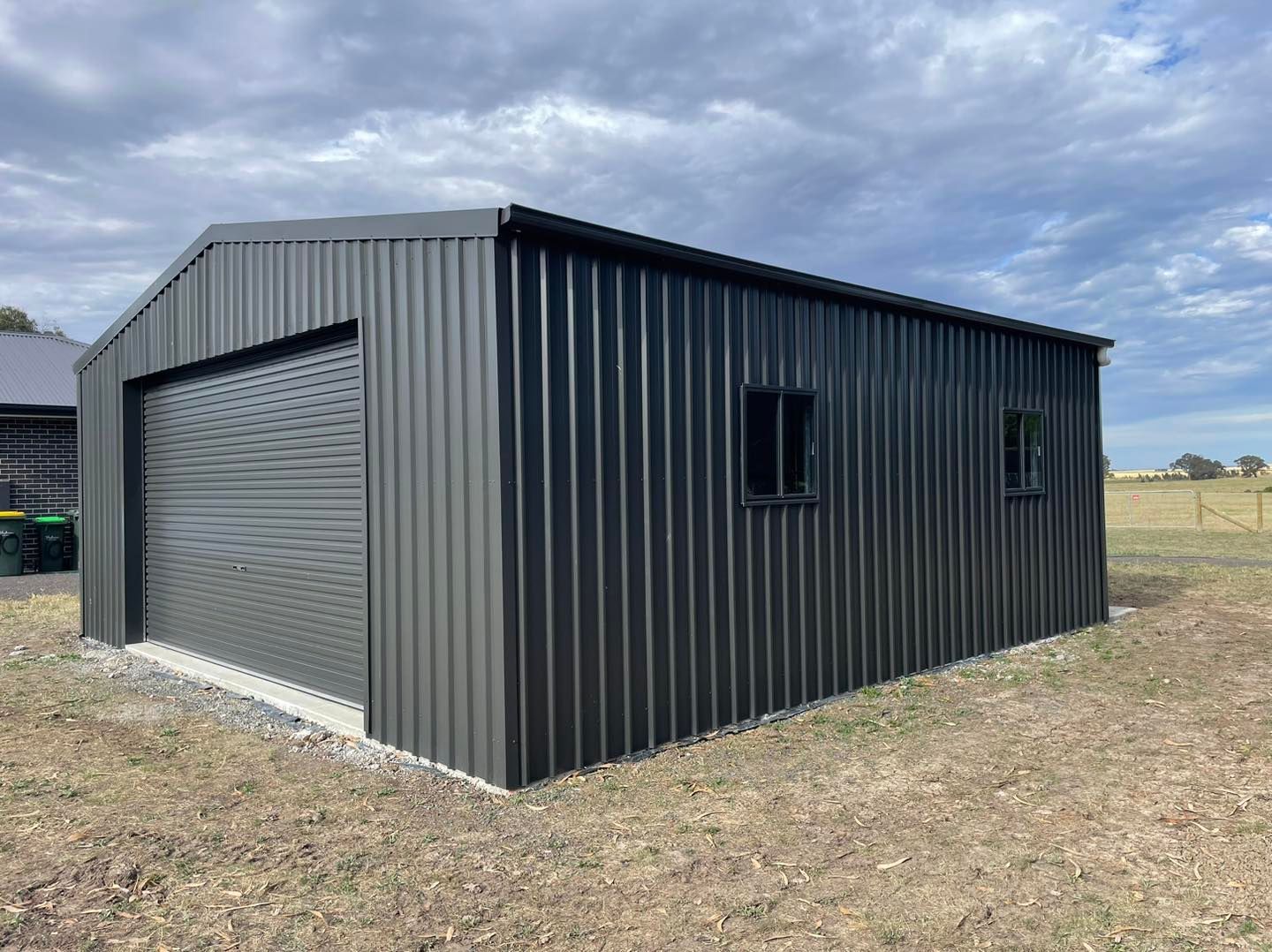 Dark gray metal shed with a roller door and two small windows on a gravel base under a cloudy sky.