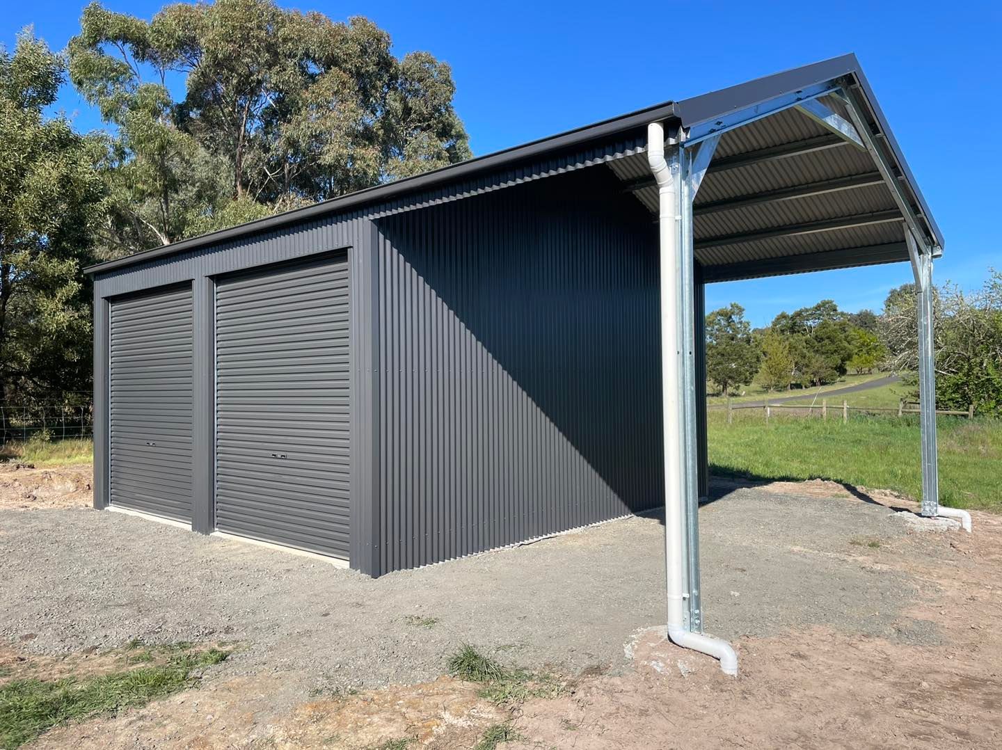 Dark gray metal shed with two garage doors and a carport in a gravel lot under a blue sky.