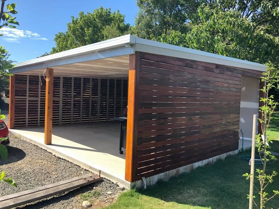 Wooden-sided outdoor structure with concrete floor, light roof, and wooden support beams on a lawn.