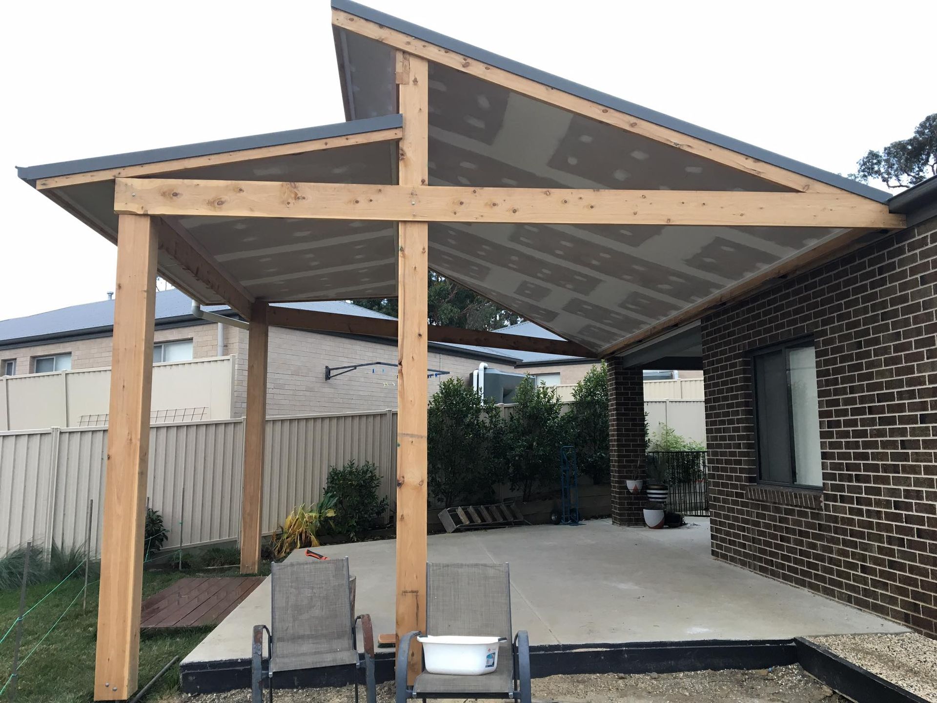 Wooden pergola attached to a brick house, over a concrete patio. Two chairs are in front.