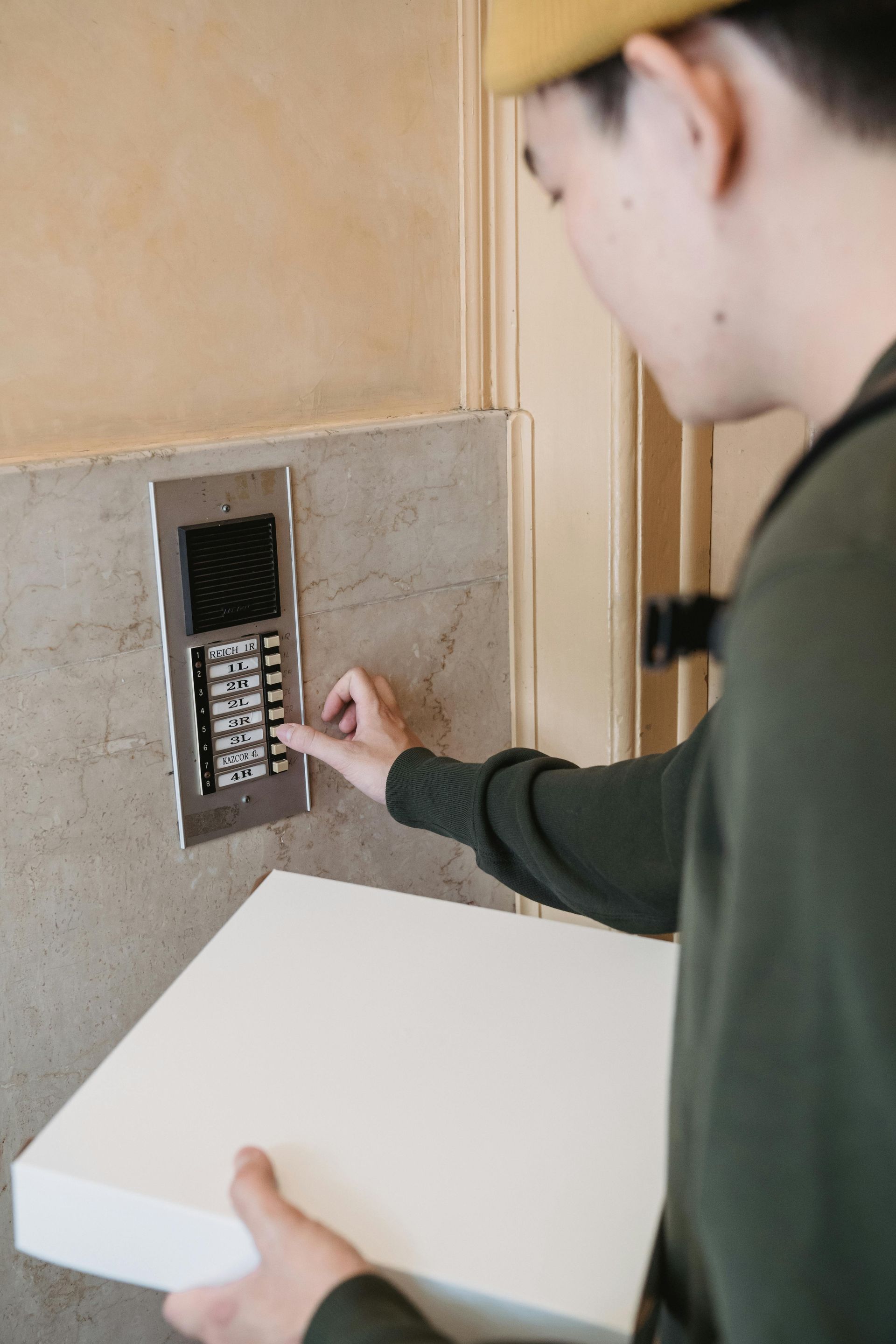 Person pressing buttons on an intercom with a pizza box, likely delivering to an apartment.