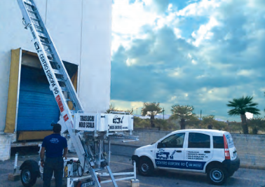 Worker and utility truck beside a building, using an extended ladder near a blue shutter under cloudy sky