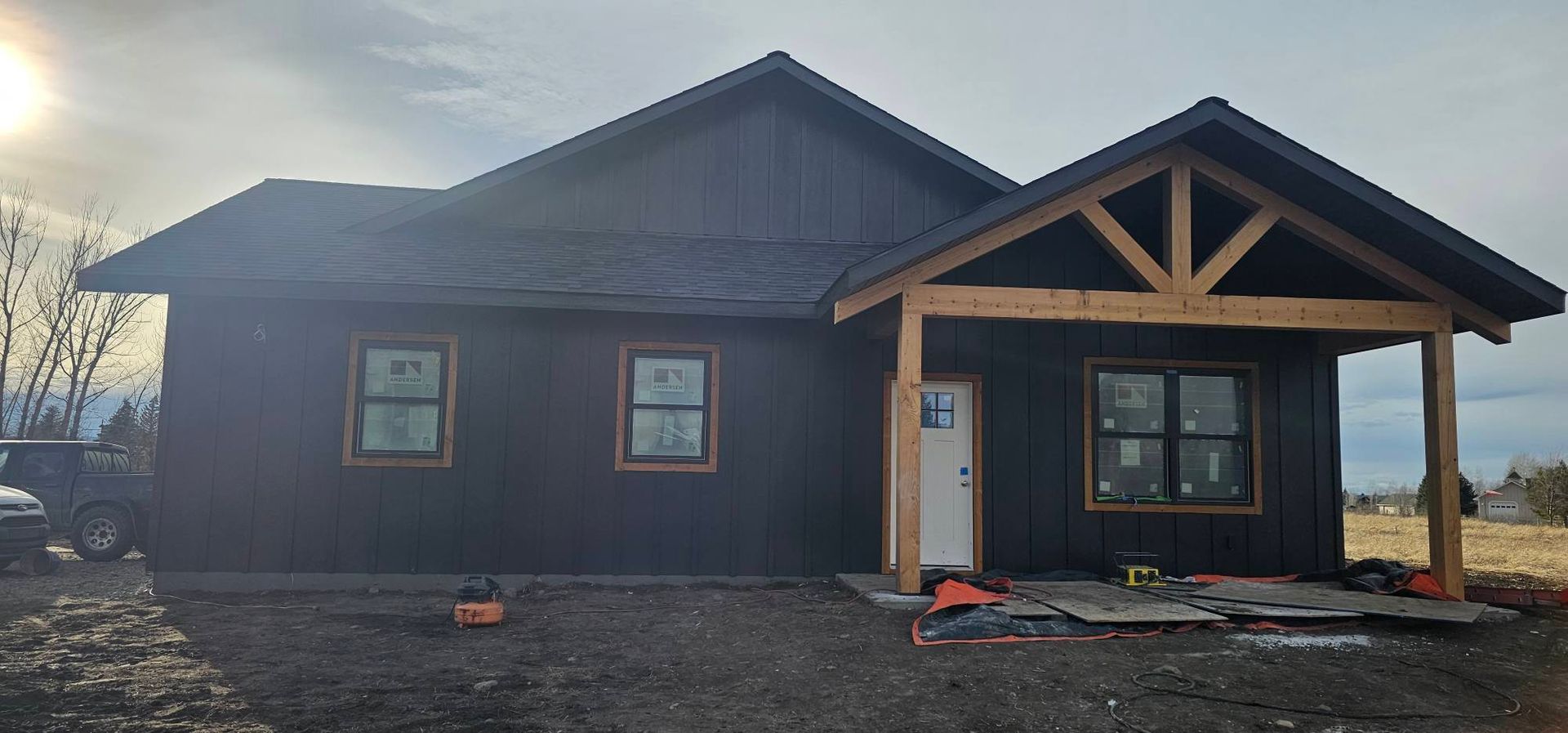 A dark-sided house under construction with a timber-framed front porch and white door against a cloudy sky.