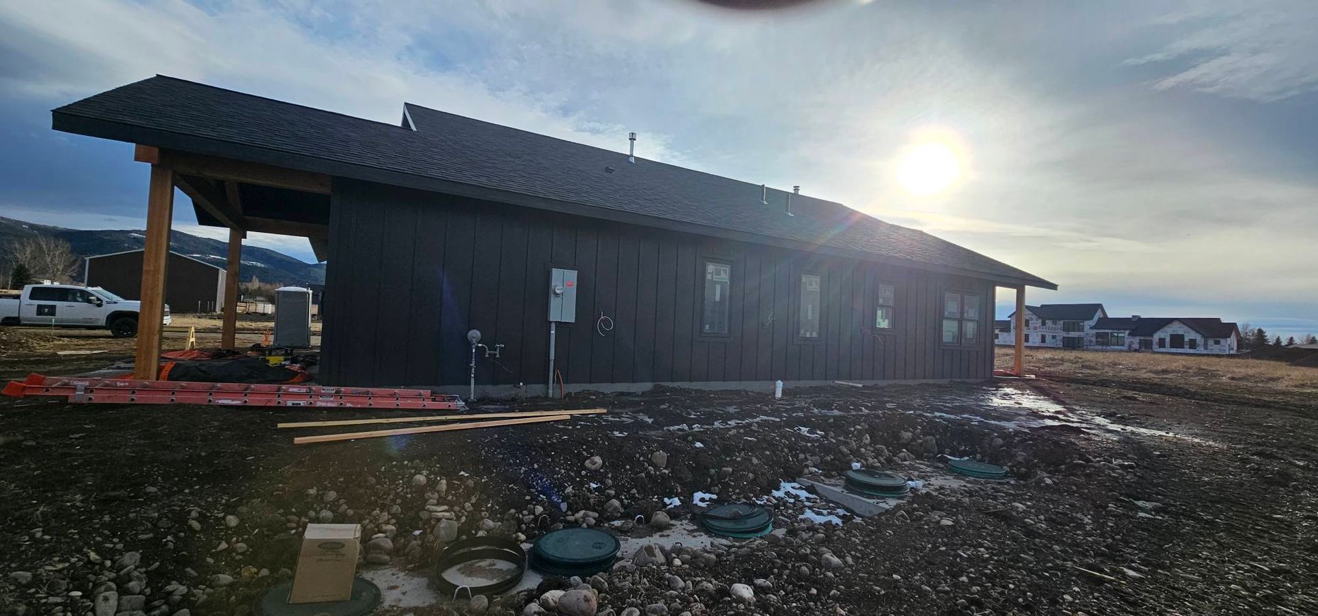 A new house under construction with dark wood siding and a pitched roof, set in a rural area under a bright, sunny sky.