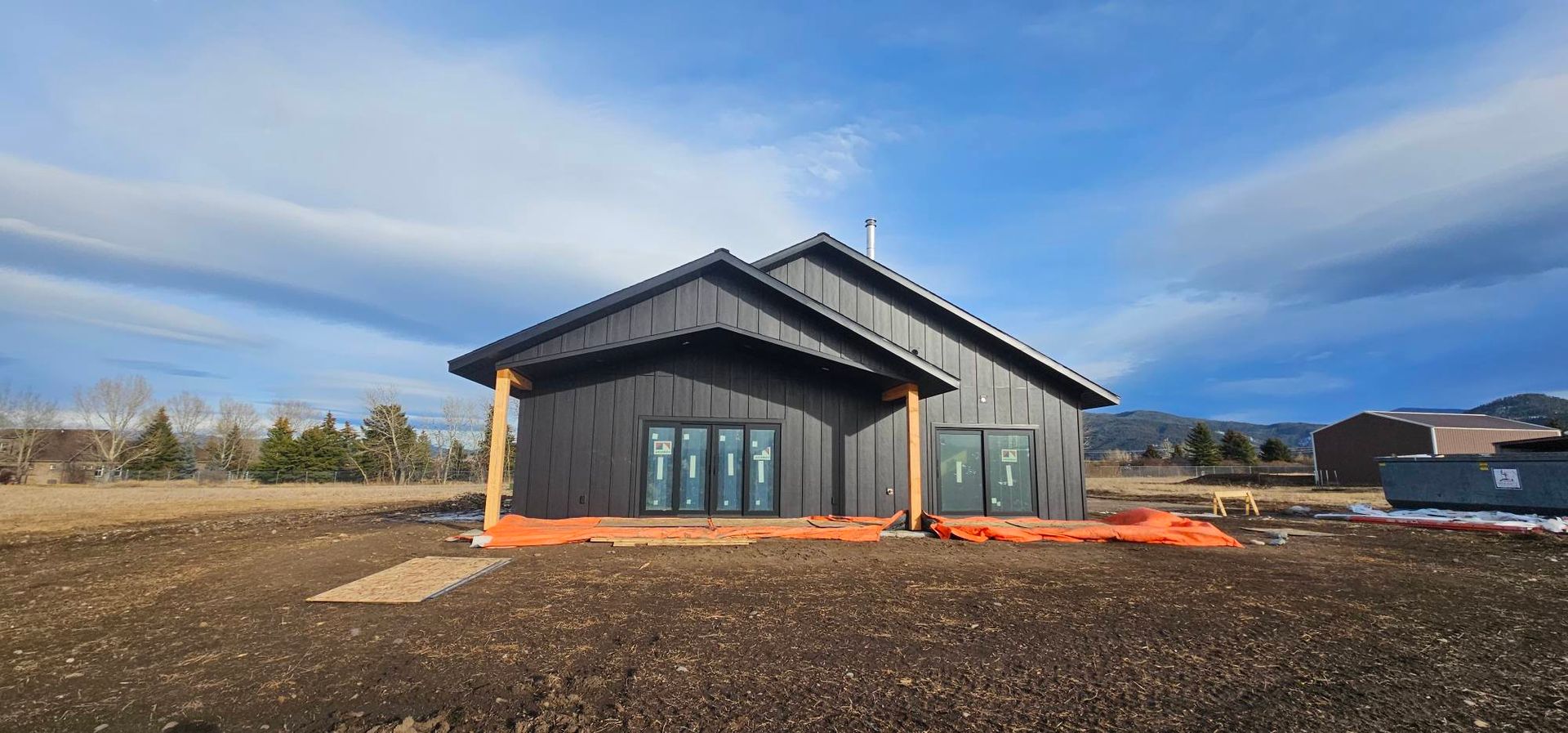 A modern, black-sided house under construction with large windows, wooden pillars, and a dirt yard under a blue sky.