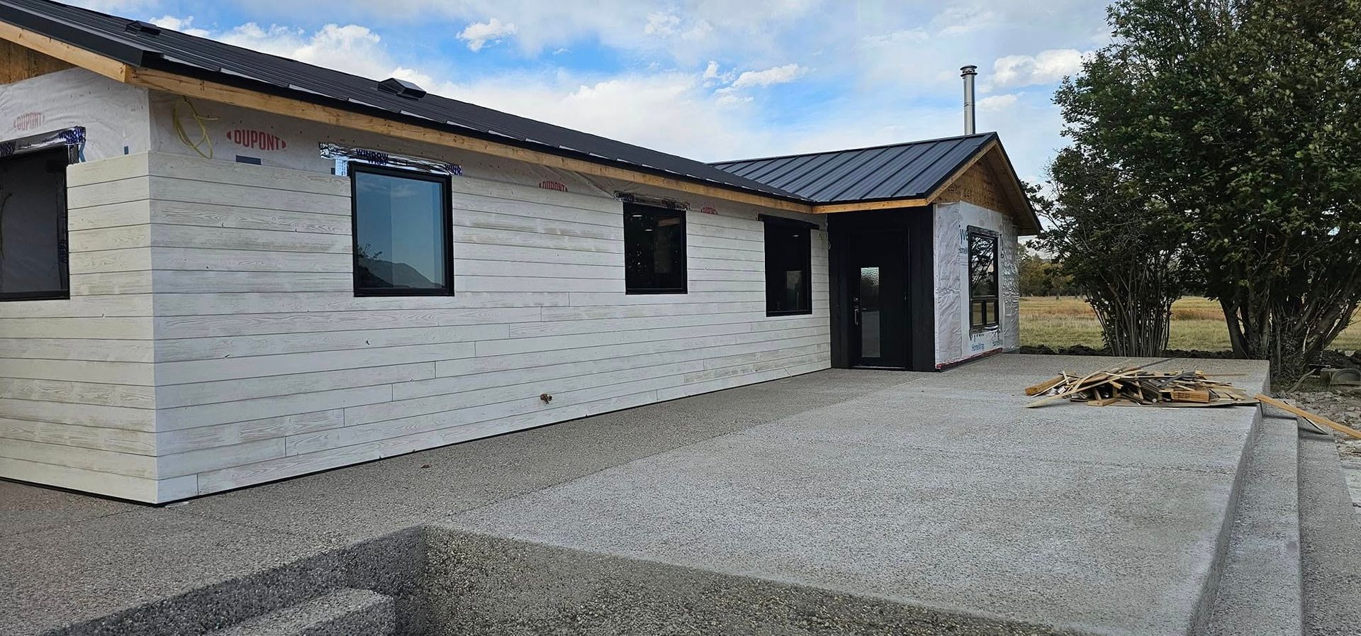 A modern, L-shaped home under construction with white exterior house wrap, dark window frames, and a concrete patio.