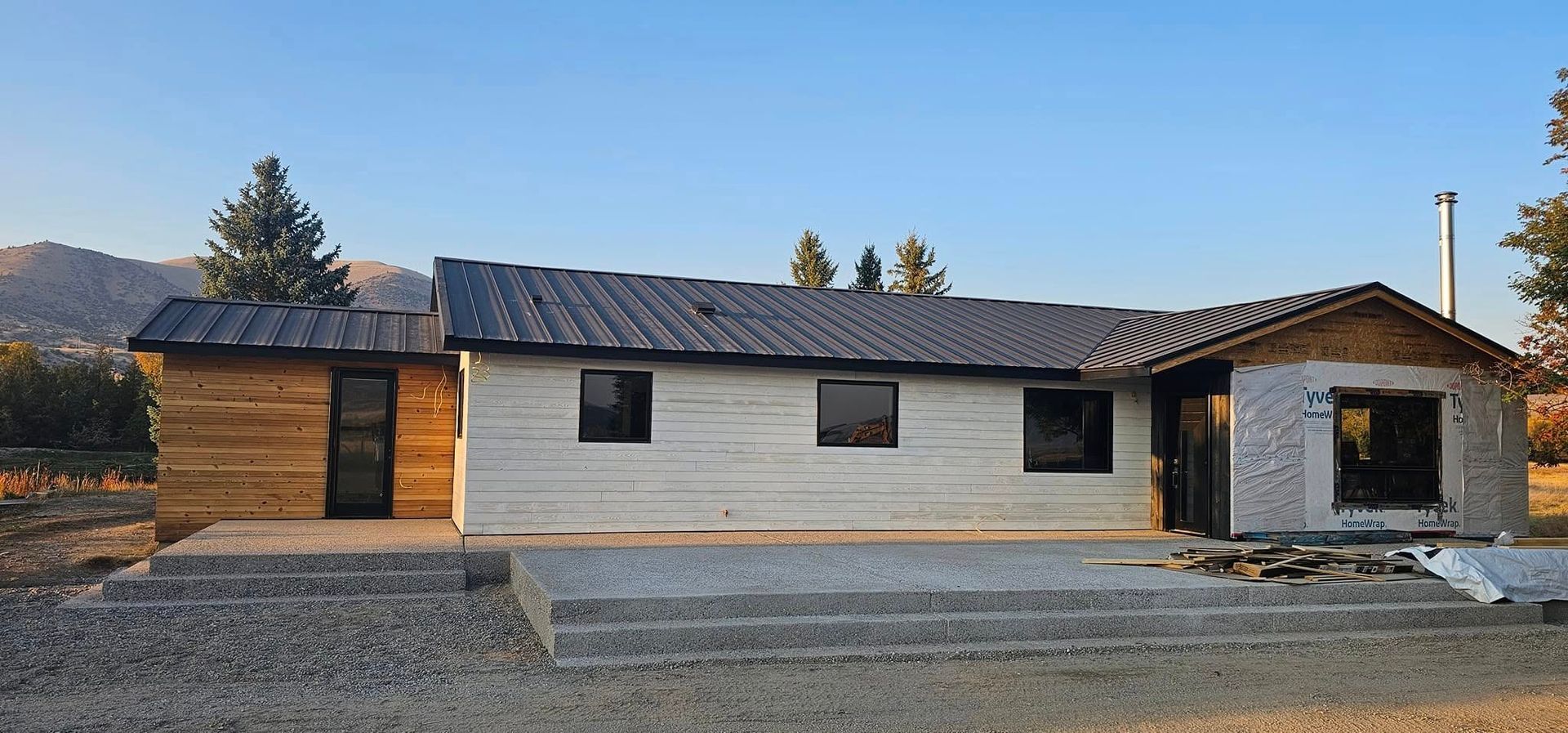 A single-story house under construction with white siding, wood accents, a dark metal roof, and a gravel patio area.