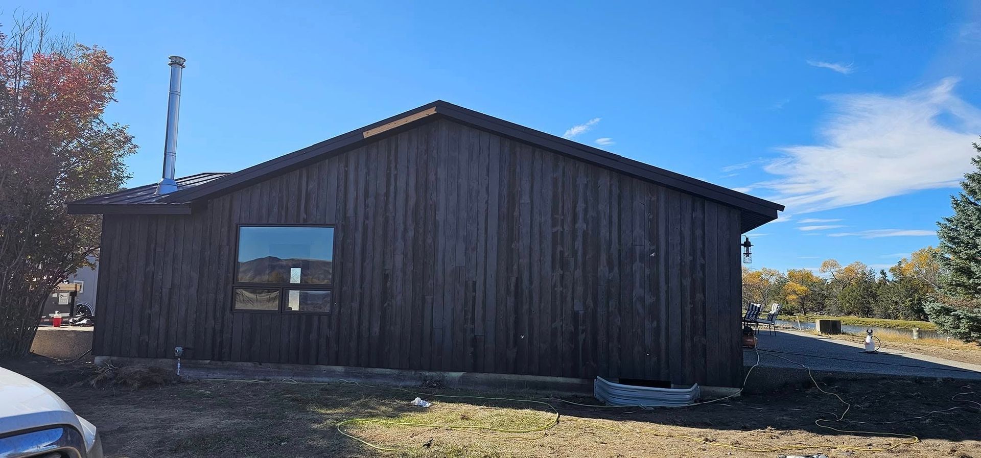 A side view of a dark brown, vertical-siding house with a single window and metal chimney against a blue sky.