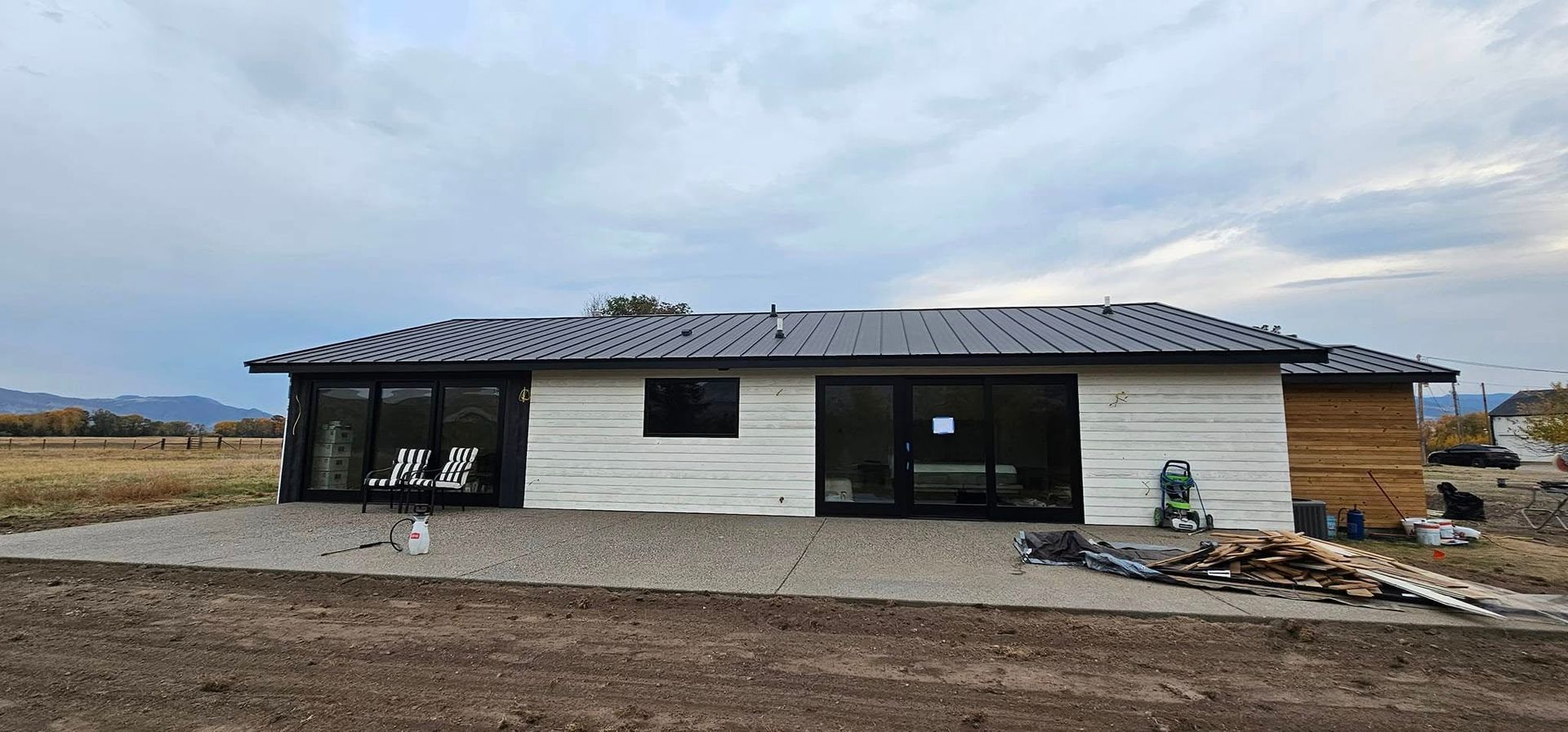 A modern one-story home with white walls and a dark metal roof, set against a rural landscape under a cloudy sky.