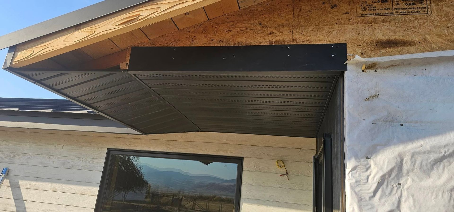 Black soffit and fascia installed under the unfinished roof overhang of a house with white exterior house wrap.