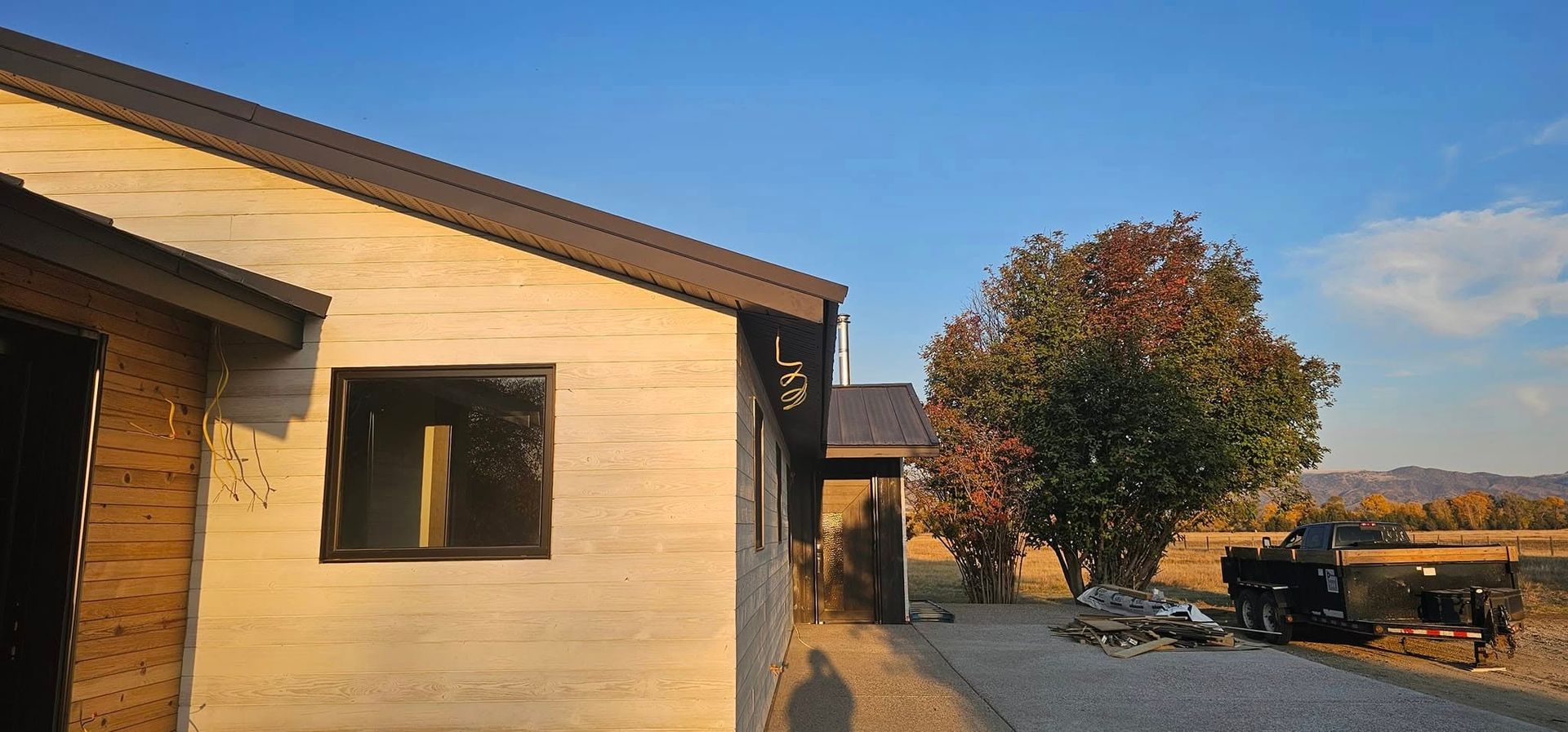 A light-colored house with a dark roof stands next to a driveway and a large, full tree under a clear blue sky.