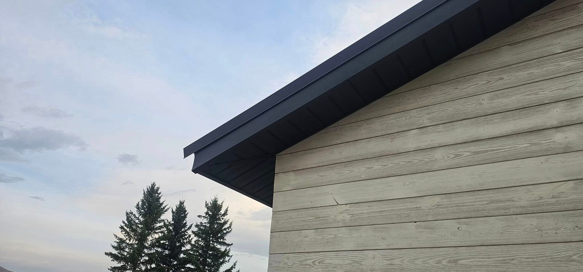 The corner of a light-colored building with horizontal textured siding under a dark roof eave against a pale sky.