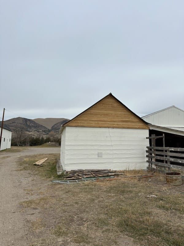 A white-walled shed with a wooden gable roof sits on a dirt road, with mountains in the background under a cloudy sky.