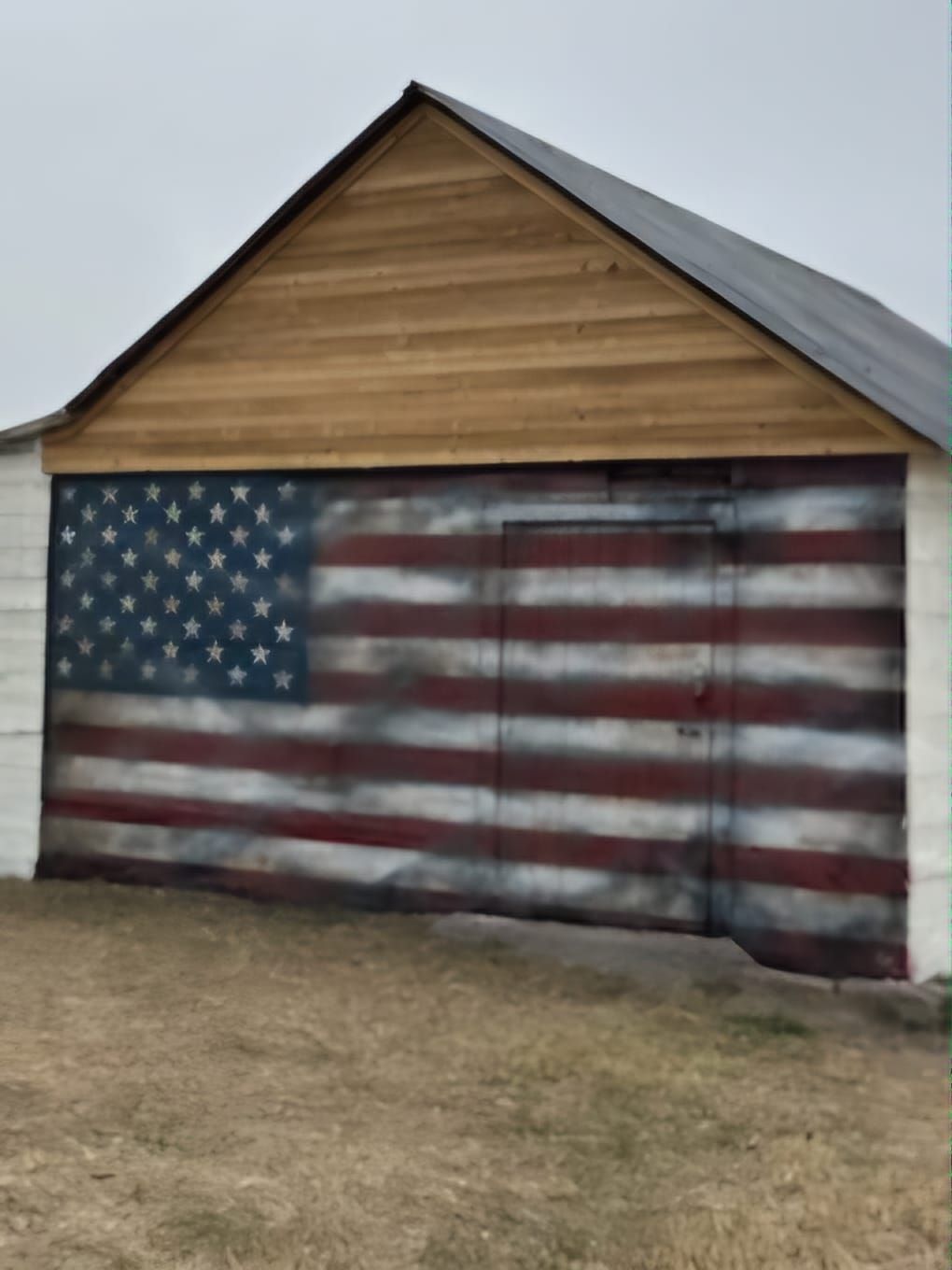 A shed with a large, weathered American flag painted on its door and a light-colored wooden gable above it.