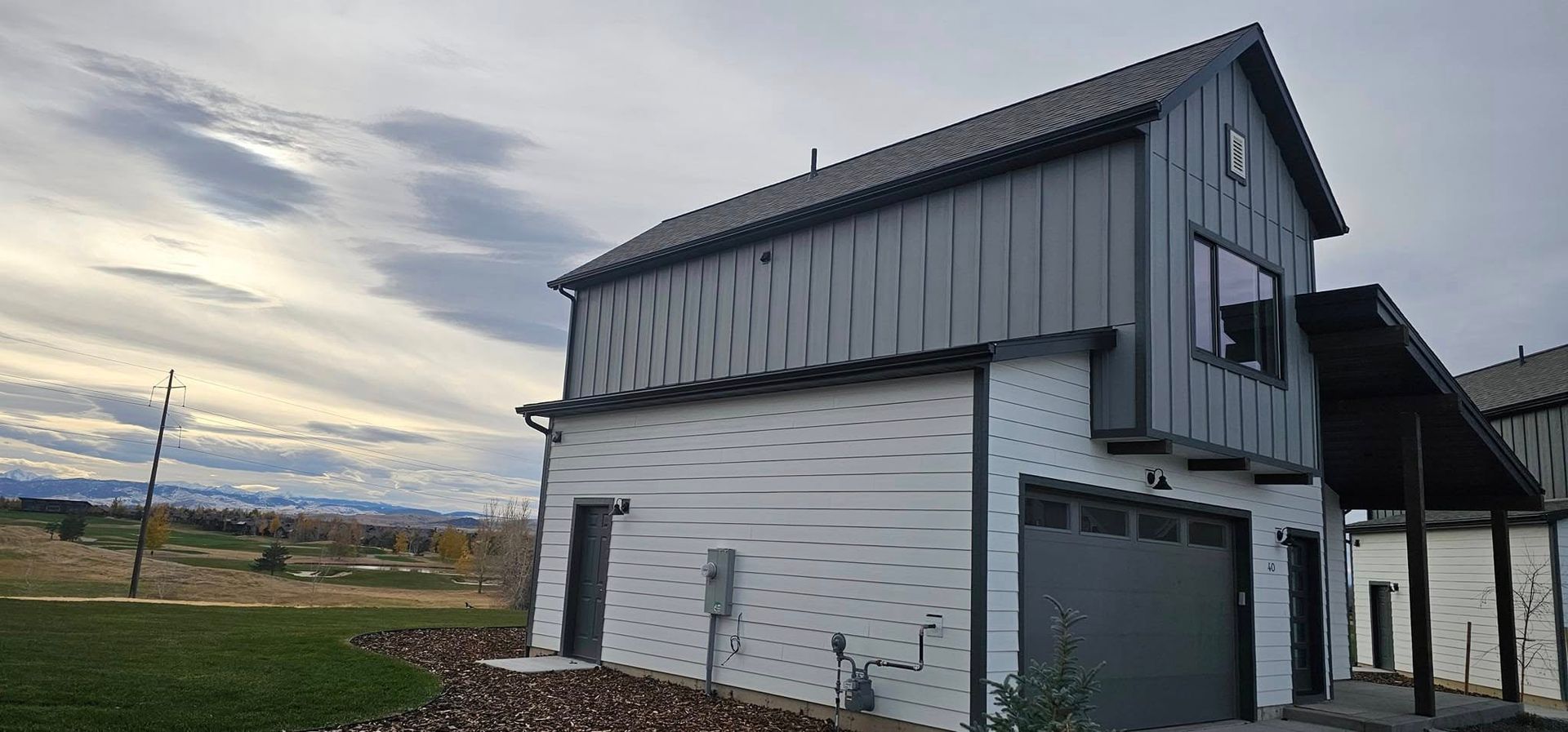 A modern two-story building with white brick on the ground floor and gray vertical metal siding above, set in a field.