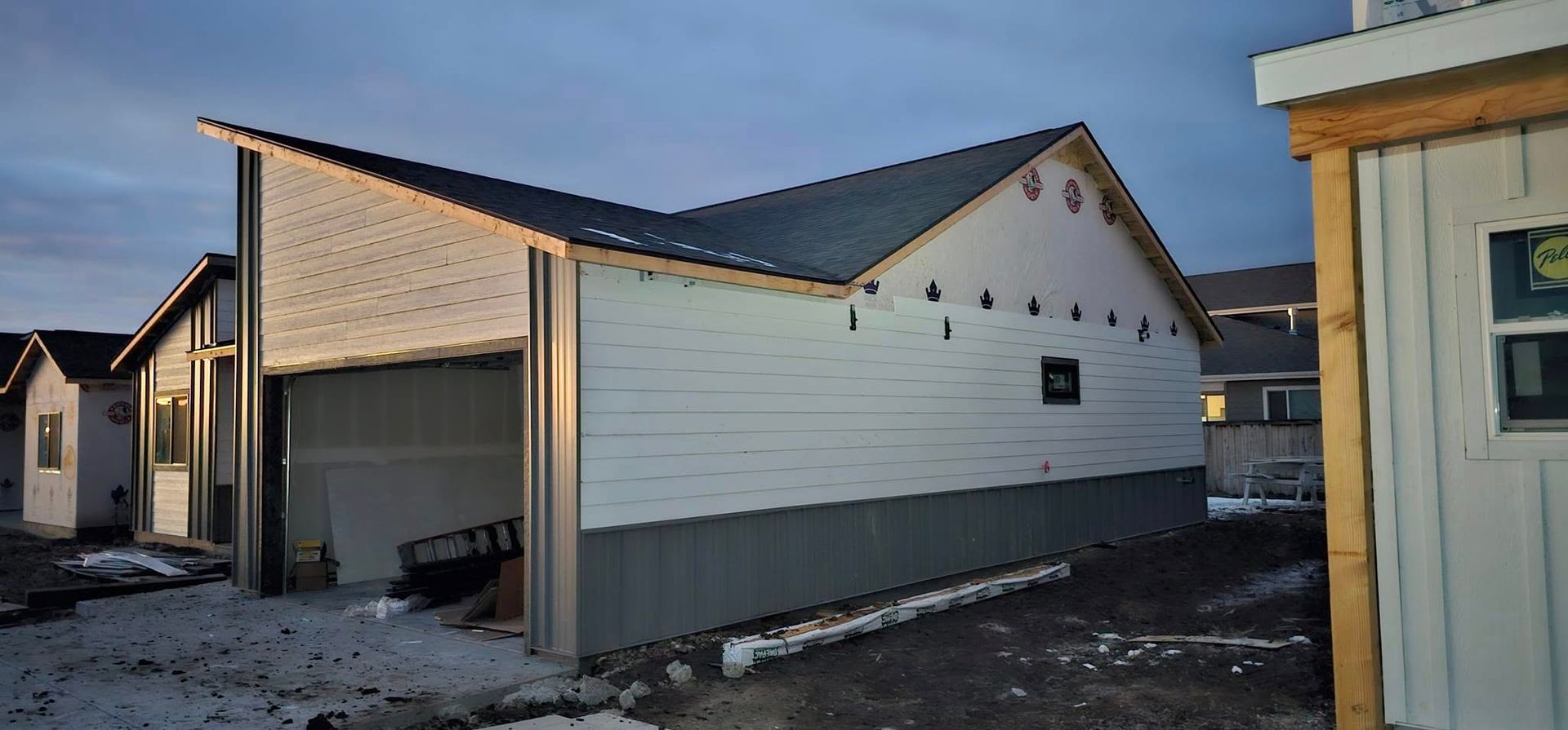 A half-finished house under construction at dusk, featuring metal siding, a garage opening, and a snowy yard.