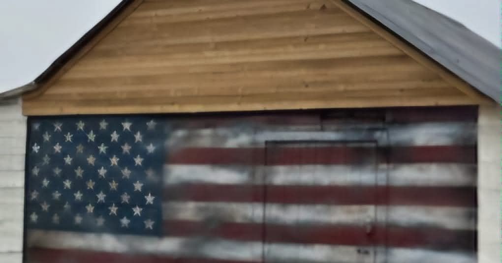 A shed wall painted with a weathered, distressed American flag beneath a natural wood gable.
