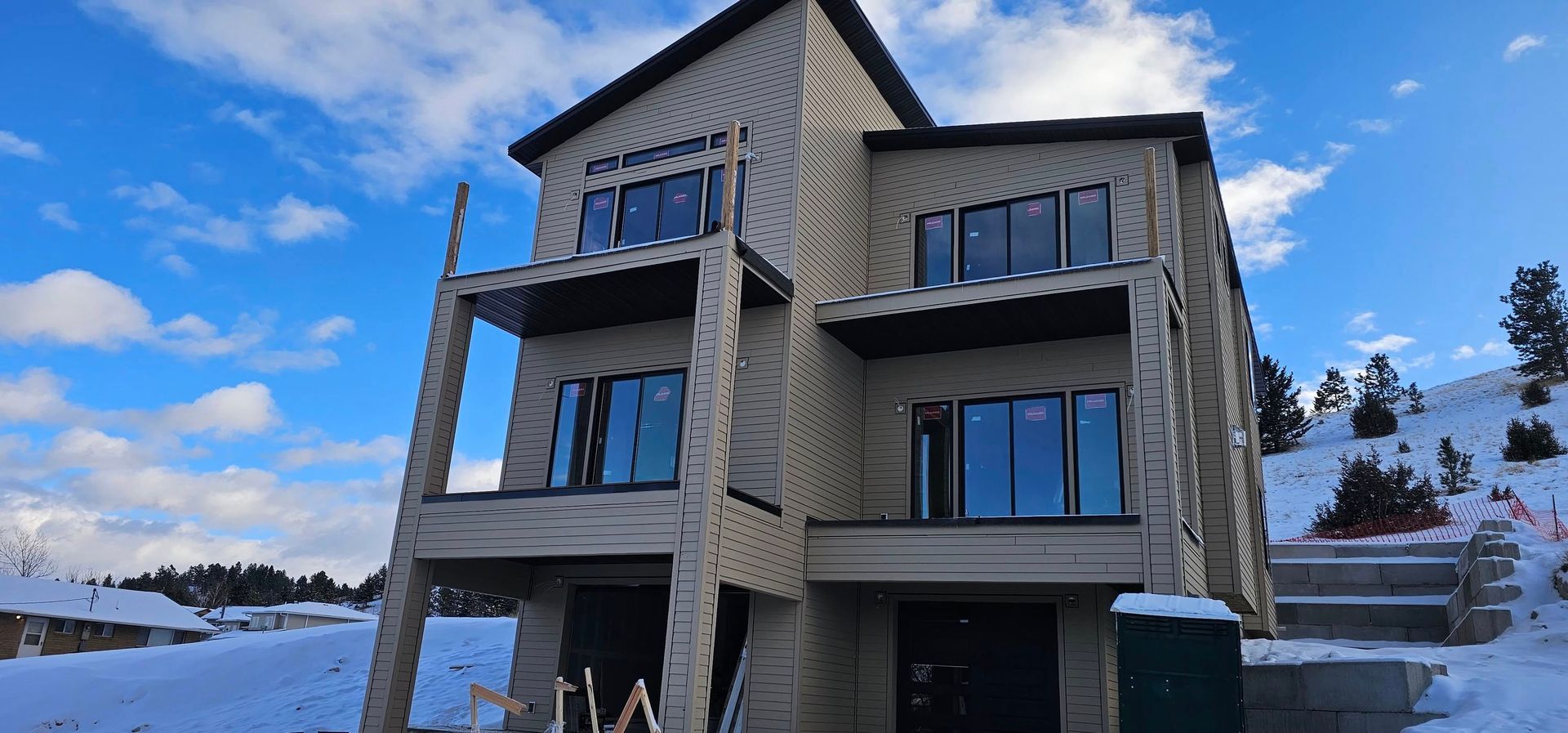 A multi-story house under construction with tan siding and balconies, set against a snow-covered landscape and blue sky.