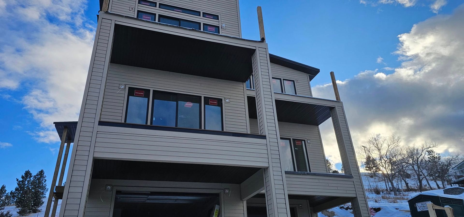 A modern three-story house with light-colored textured siding and balconies, set against a bright, cloudy sky and snow.