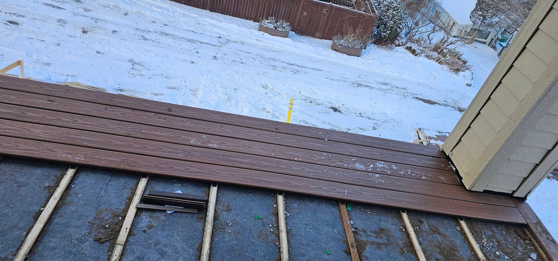 A close-up of a partially installed roof, showing wooden joists, black underlayment, and a row of brown metal roof panels.