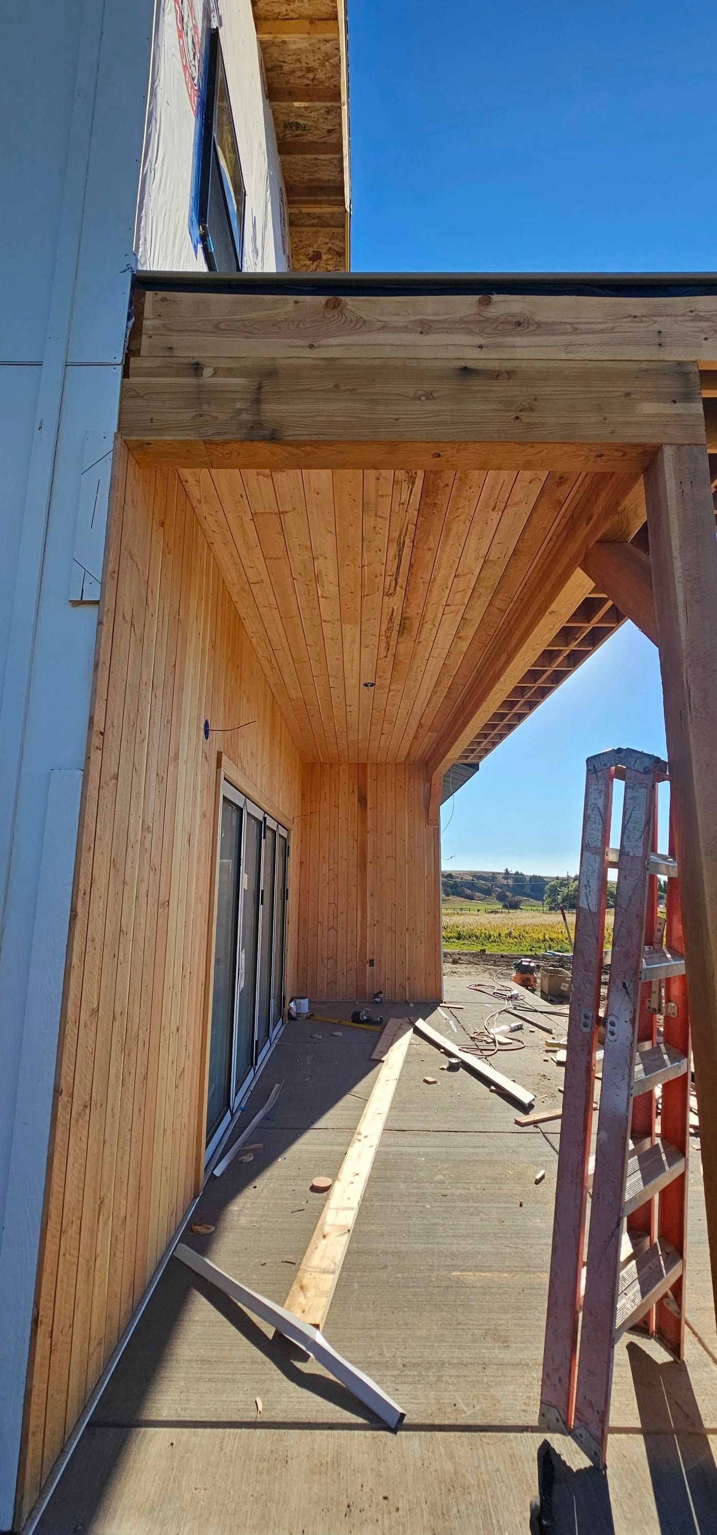 A low-angle view of a building under construction featuring a wooden overhang and a tall ladder on a concrete walkway.