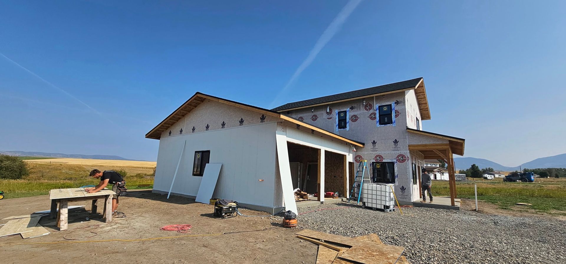 Construction site with a partially built house featuring white walls and exposed insulation, with a worker at a table.